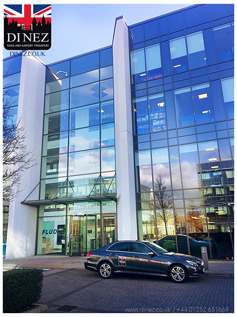 FLUOR Office in Farnborough with an executive Mercedes taxi parked in front of the glass building. The log of Dinez Taxis and Airport Transfers is on the upper left side that states 'DINEZ', a union jack flag with a silhouette of Farnborough Town. Call ☎️01252 265051☎️ for taxi to, from and in Fluor, Farnborough